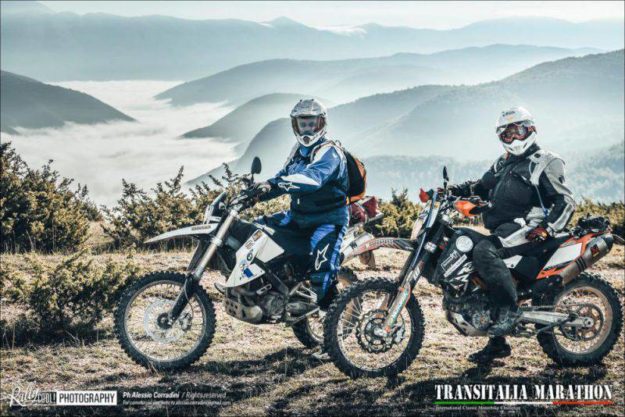 Due uomini in moto con dietro un panorama di montagne e fiumi
